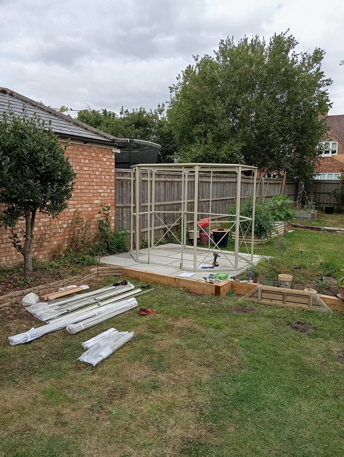 Greenhouse kit parts laid out on the lawn with the wall frame going up on a paved base
